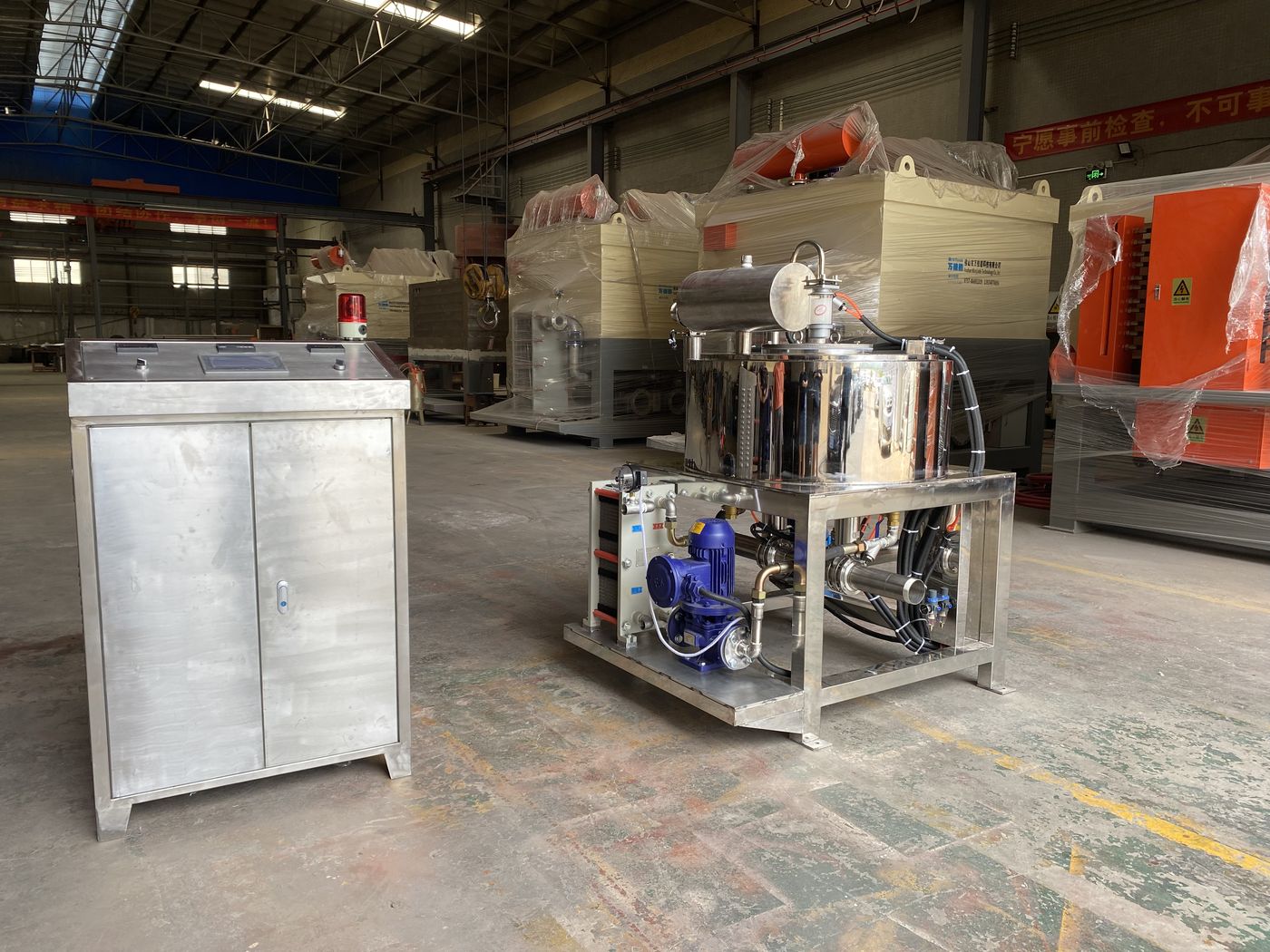Stainless-steel slurry separator next to a stainless-steel control cabinet in the PTMS workshop, with more machines wrapped for shipping behind them